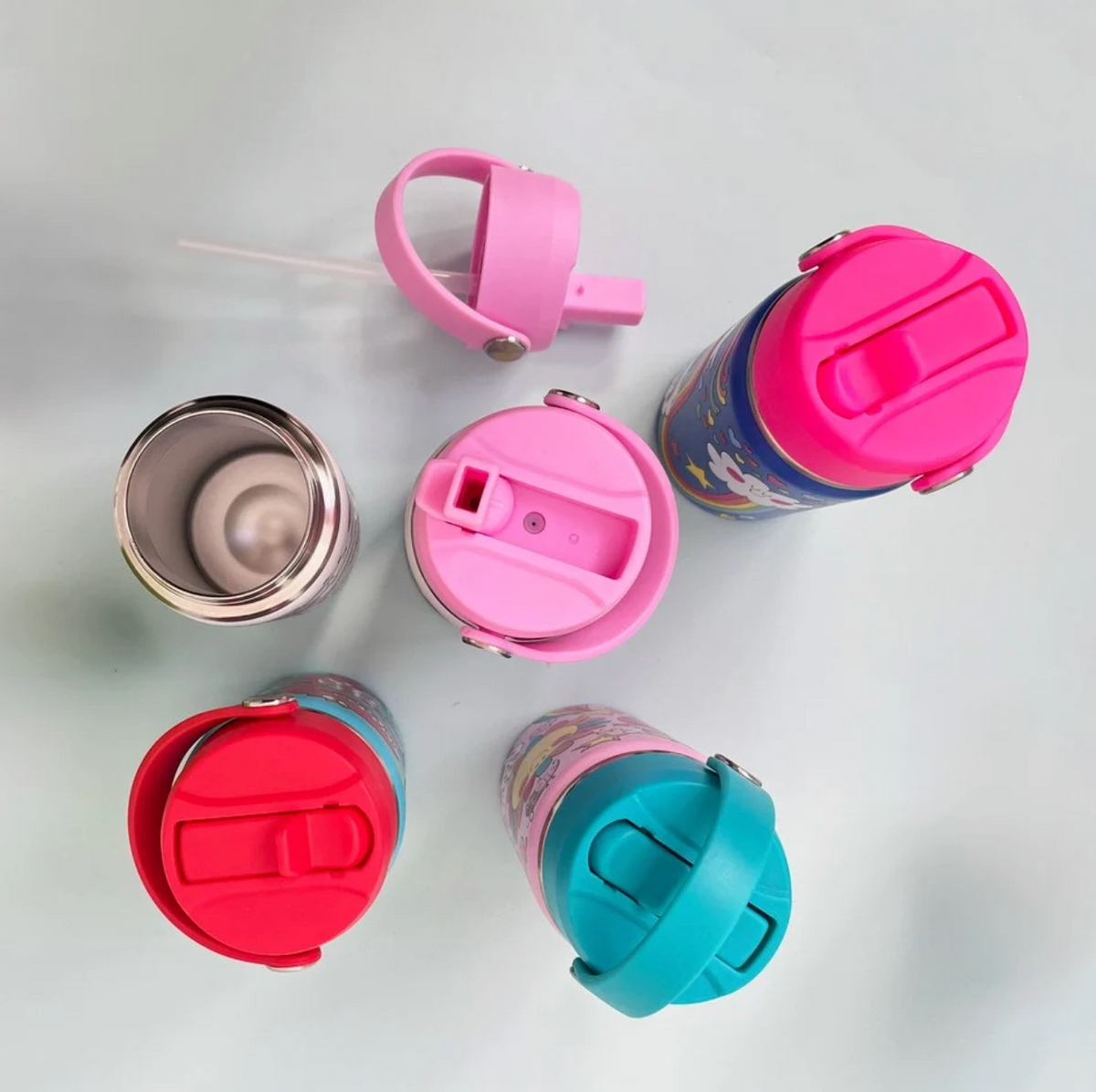 Overhead Rachel Ellen bottle range on pale blue: open stainless steel interior at left, detached pink lid and long clear straw above, raised pink spout in centre, closed fuchsia lid at upper right, red Dinosaur lid at lower left and turquoise lid with handle across it at lower right.