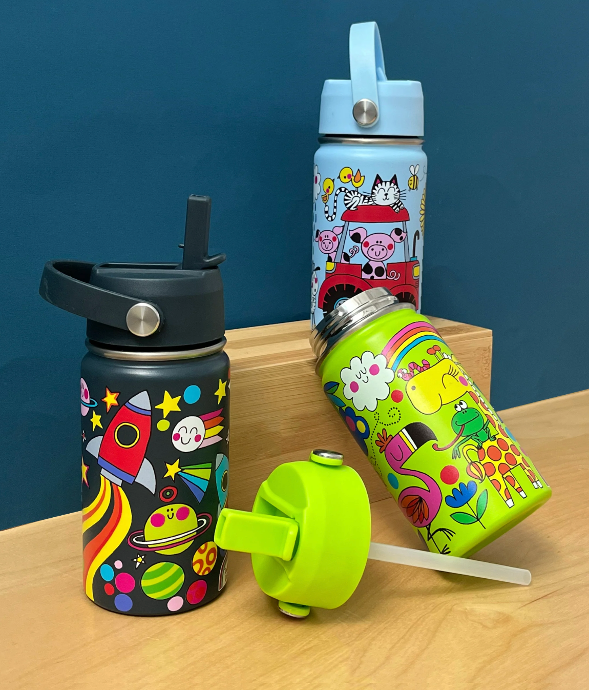 Rachel Ellen Space dark navy bottle at left on wooden tabletop against deep teal background, with raised straw, red rocket and colourful planets; pale blue Farm bottle on wooden riser behind, open lime-green Jungle bottle leaning right and detached green lid with clear straw in front.