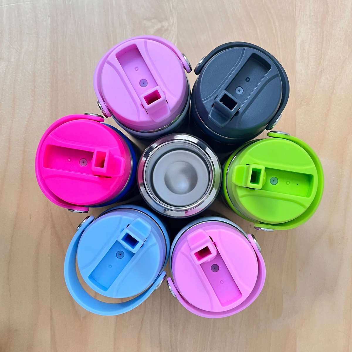 Overhead six Rachel Ellen bottles arranged around an open stainless steel bottle on wood; raised straw lids in lilac at upper left, dark navy Space at upper right, lime green at right, pale pink below right, sky blue below left and fuchsia at left, with silver handle pivots.