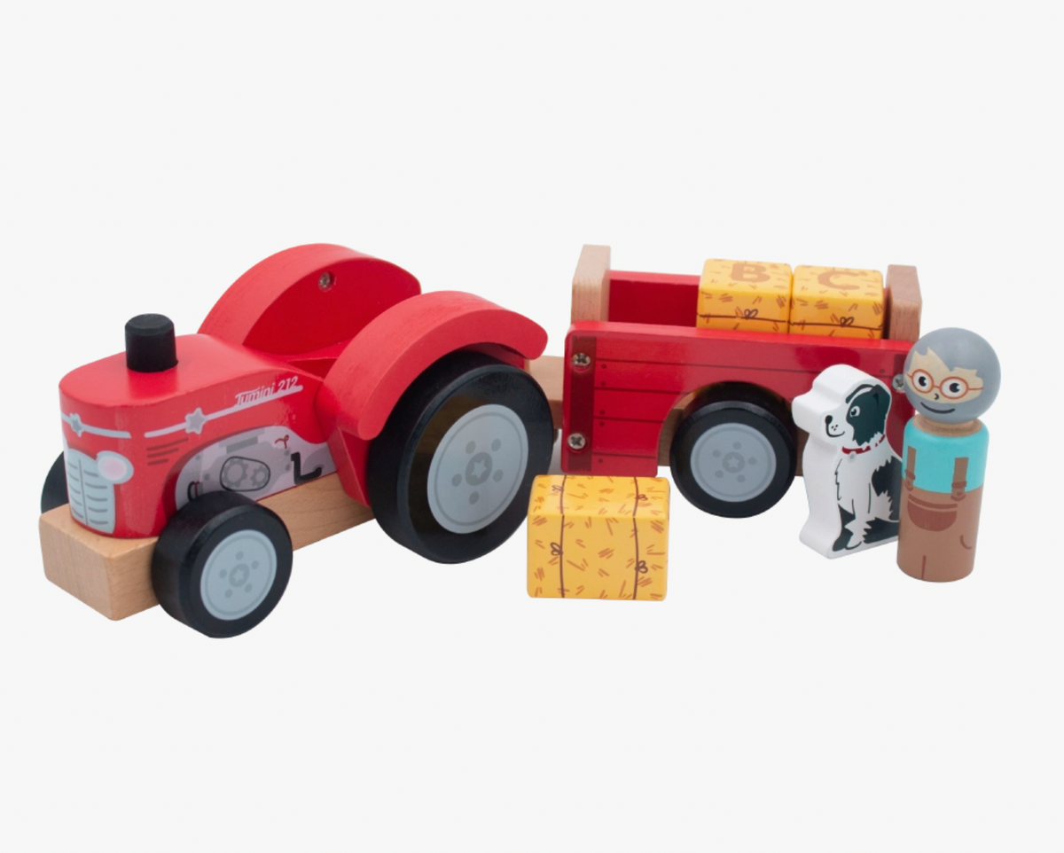 Jumini red wooden farm tractor and trailer on white background, with black wheels and grey hubs, natural wood chassis, black exhaust and printed silver engine details; two yellow hay bales in trailer, third in front, black-and-white dog with red collar and grey-haired farmer in glasses and turquoise top at right.