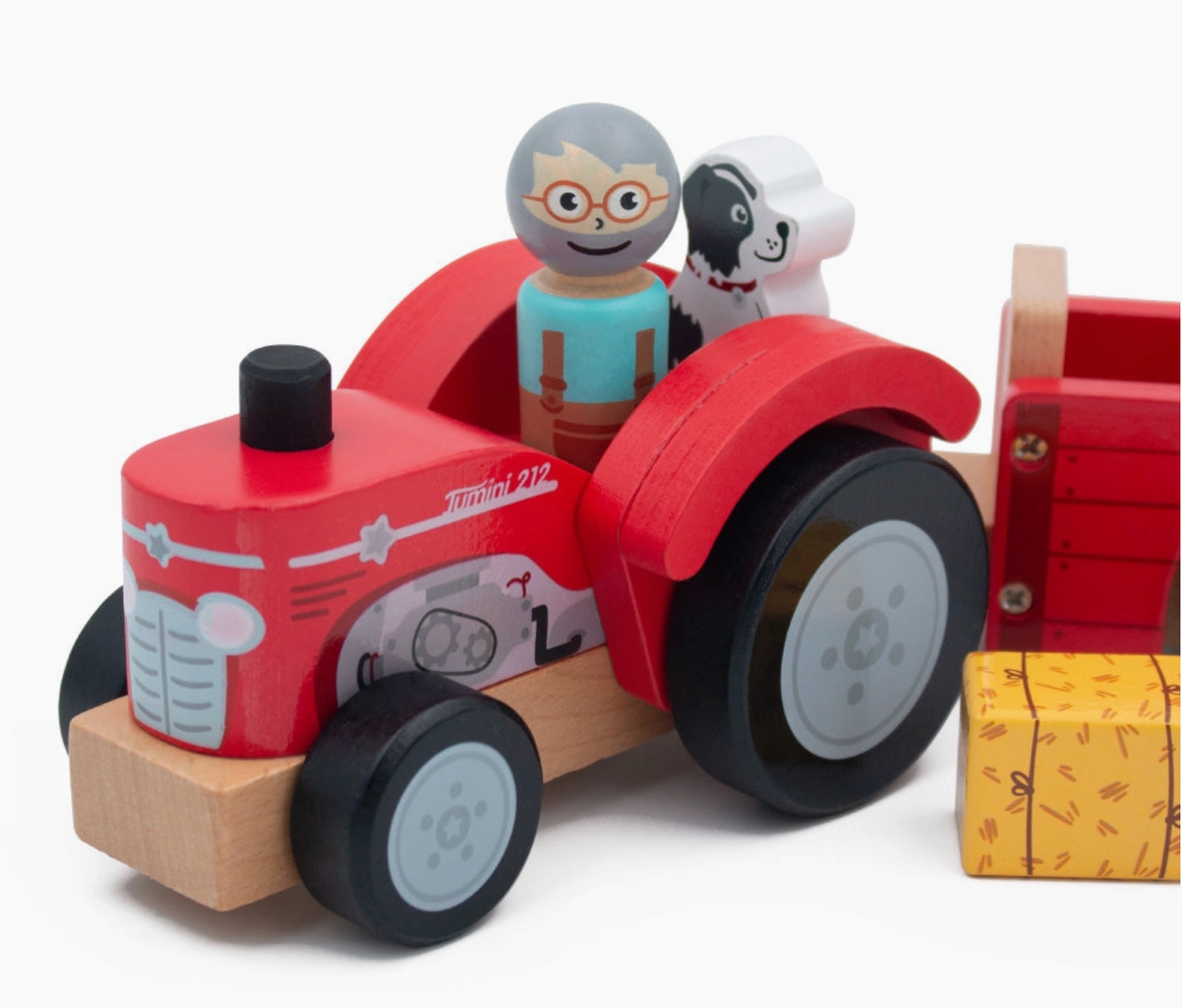 Close-up of Jumini red wooden tractor with smiling grey-haired farmer in orange-rimmed glasses, turquoise shirt and brown dungarees beside black-and-white dog; curved red wheel arches, black wheels with grey hubs, printed headlights, grille, engine and Jumini 212 marking; trailer and yellow bale cropped at right.