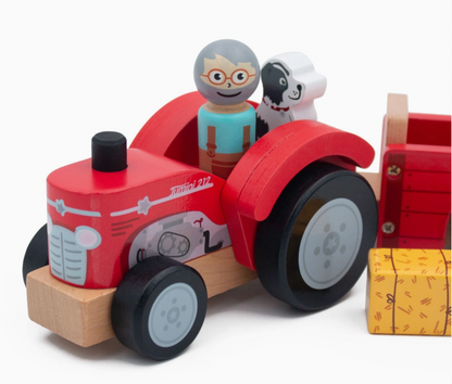 Close-up of Jumini red wooden tractor with smiling grey-haired farmer in orange-rimmed glasses, turquoise shirt and brown dungarees beside black-and-white dog; curved red wheel arches, black wheels with grey hubs, printed headlights, grille, engine and Jumini 212 marking; trailer and yellow bale cropped at right.