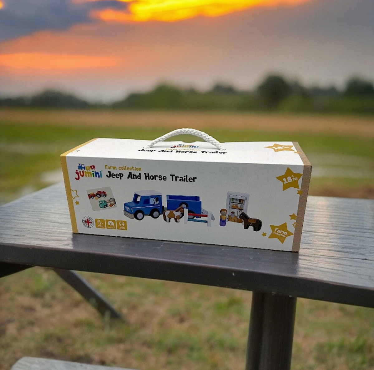 Jumini Jeep And Horse Trailer white gift box with rope handle on outdoor wooden picnic table at sunset; front shows multicoloured Jumini logo, Farm collection title, playset illustration, yellow star marked 18m+, Union Jack emblem and small yellow feature icons, with fields and orange sky behind.