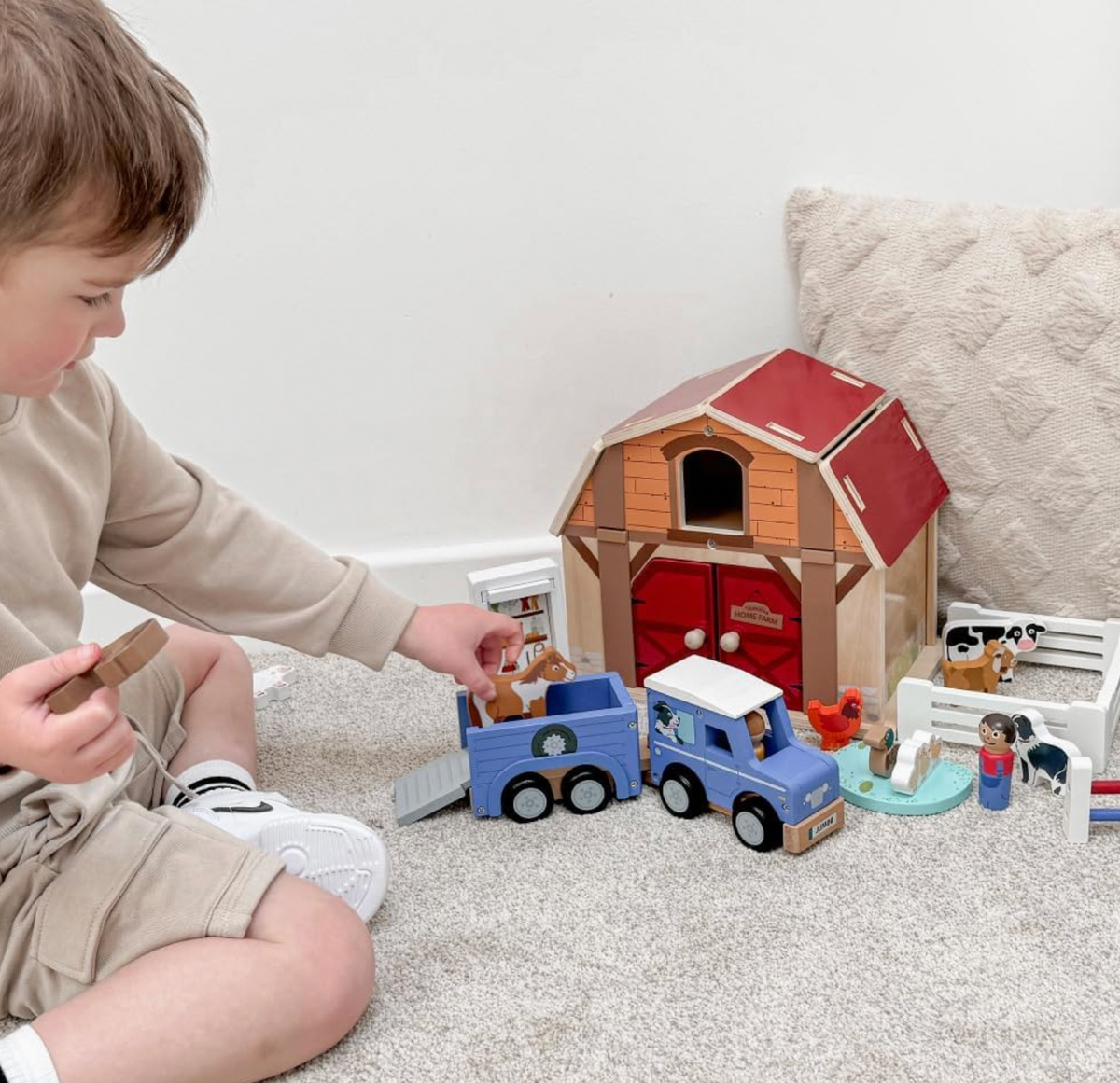 Child seated on cream carpet placing a wooden horse into Jumini blue horse trailer beside matching jeep, while holding another horse; detached roof stands as tack storage behind. Larger red-roofed barn, fenced animals, figures and farm accessories create a wider farm scene.