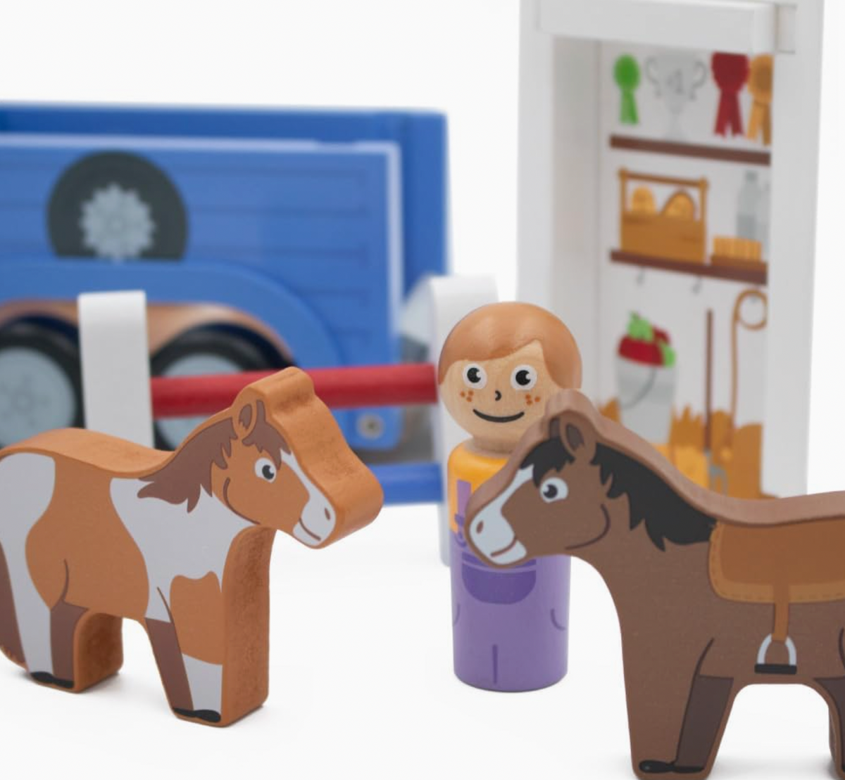 Close-up of Jumini chestnut-and-white wooden horse at left and brown horse with black mane, white facial blaze and printed tan saddle and stirrup at right; smiling ginger-haired peg person in yellow top and purple dungarees between them, with red-and-blue jump fence, blue trailer and illustrated tack storage behind.
