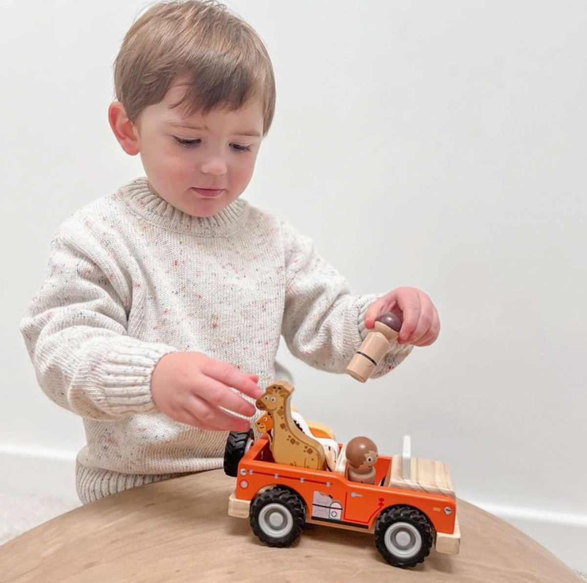 Child in cream flecked knitted jumper holding a wooden safari figure above Jumini orange jeep on a wooden table, with other hand near giraffe; zebra and lion loaded in open rear, another figure seated behind pale windscreen, black treaded wheels and printed side equipment visible.