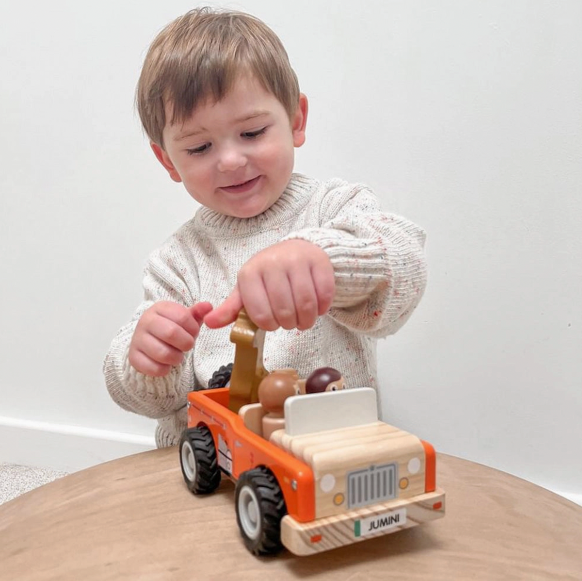 Child in cream flecked knitted jumper placing giraffe into Jumini orange safari jeep on wooden table; two figures seated behind pale windscreen, rear spare wheel visible, with natural-wood bonnet, printed grey grille, round headlights and JUMINI front plate facing camera.