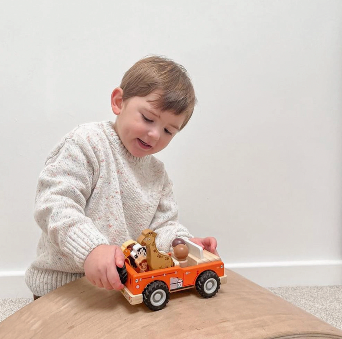 Child in cream ribbed-cuff knitted jumper holding loaded Jumini orange wooden safari jeep on tabletop; giraffe rises above lion, zebra and monkey in open rear, two figures seated in front, black treaded wheels with grey hubs and printed side equipment shown in side view.