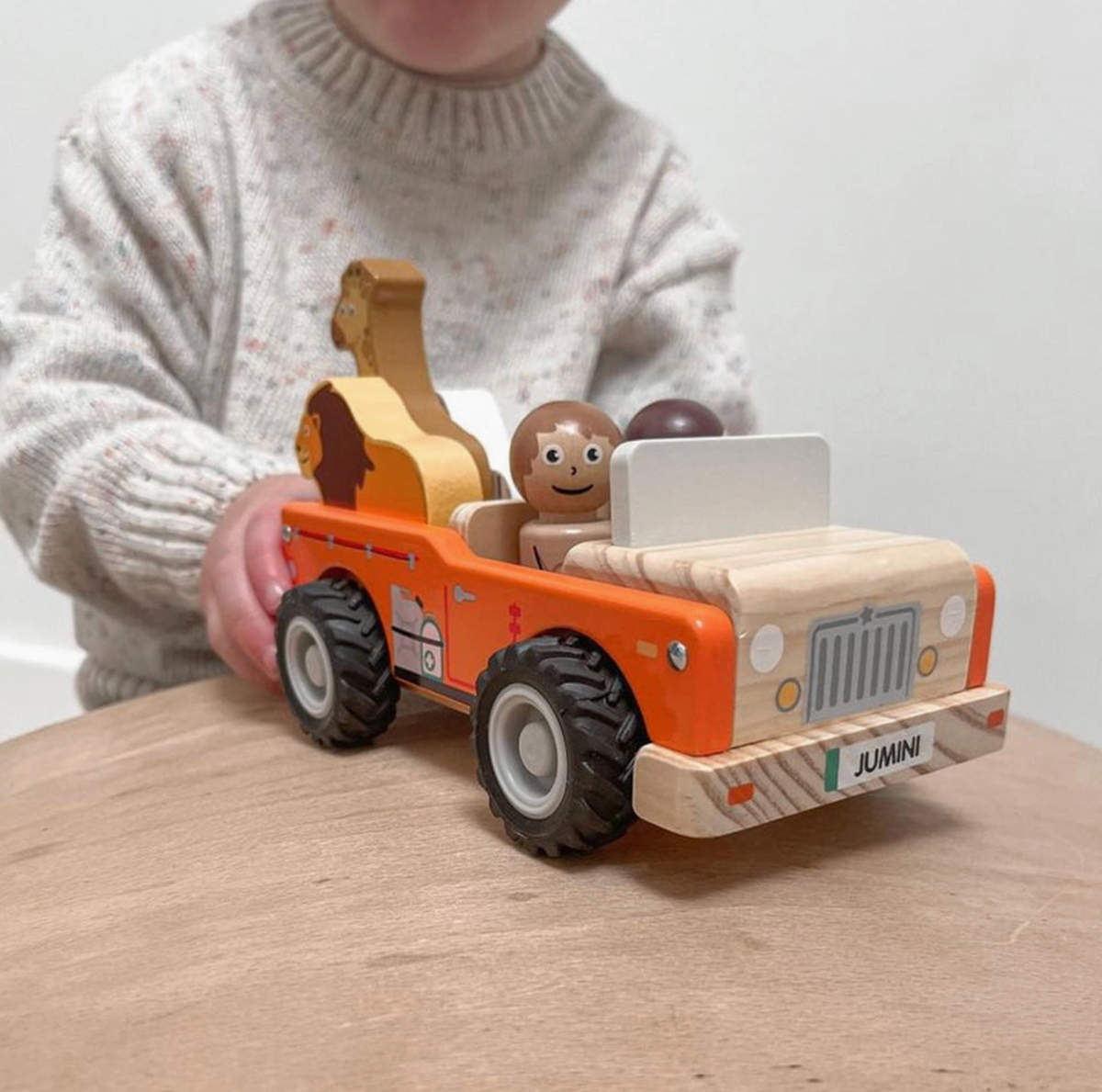 Close-up of Jumini orange safari jeep held by child on wooden table; natural-wood bonnet and bumper show wood grain, printed grille, white headlights, yellow lamps and JUMINI plate with green stripe. Black deeply treaded wheels, pale windscreen, smiling figure, lion and giraffe visible