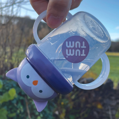 Baby sippy cup with purple lid featuring a cartoon character, held outdoors. TIPPY CUP WITH STRAW AND FLIP TOP LEAKPROOF LID - OLIVIA OWL PURPLE | TUM TUM