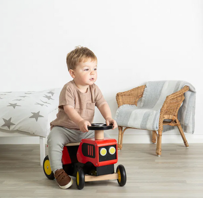 Child playing with a red toy car in a room with a couch and chair. TODDLER RIDE ON WOODEN TRACTOR - RED | BIGJIGS