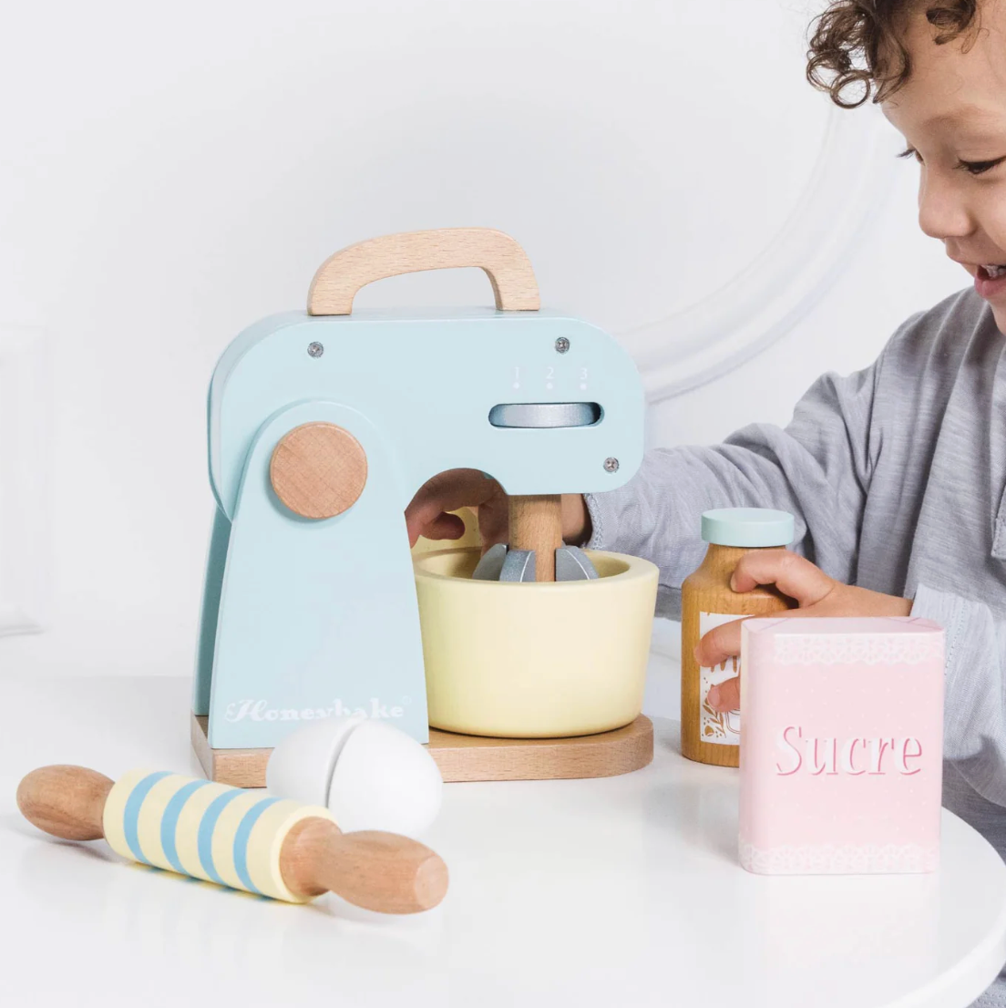 Child playing with a toy kitchen set on a white surface TOY BAKERS MIXER SET AND ACCESSORIES | LE TOY VAN