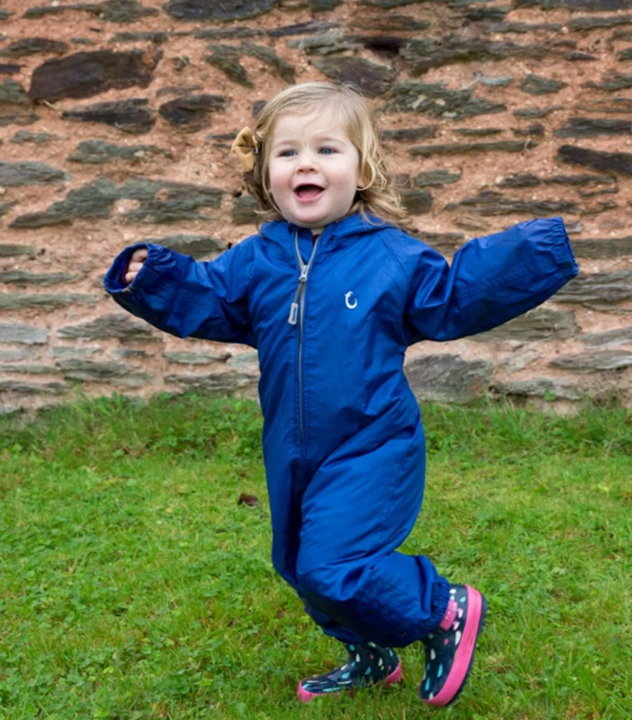 Child wearing a blue raincoat jumping in front of a stone wall. WATERPROOF TODDLER ALL-IN-ONE PACKASUITS - BLUE | HIPPYCHICK 