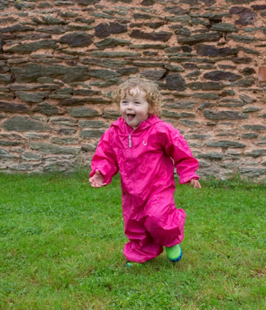 Child in a pink raincoat running on grass with a stone wall background. WATERPROOF TODDLER ALL-IN-ONE PACKASUITS - PINK | HIPPYCHICK 
