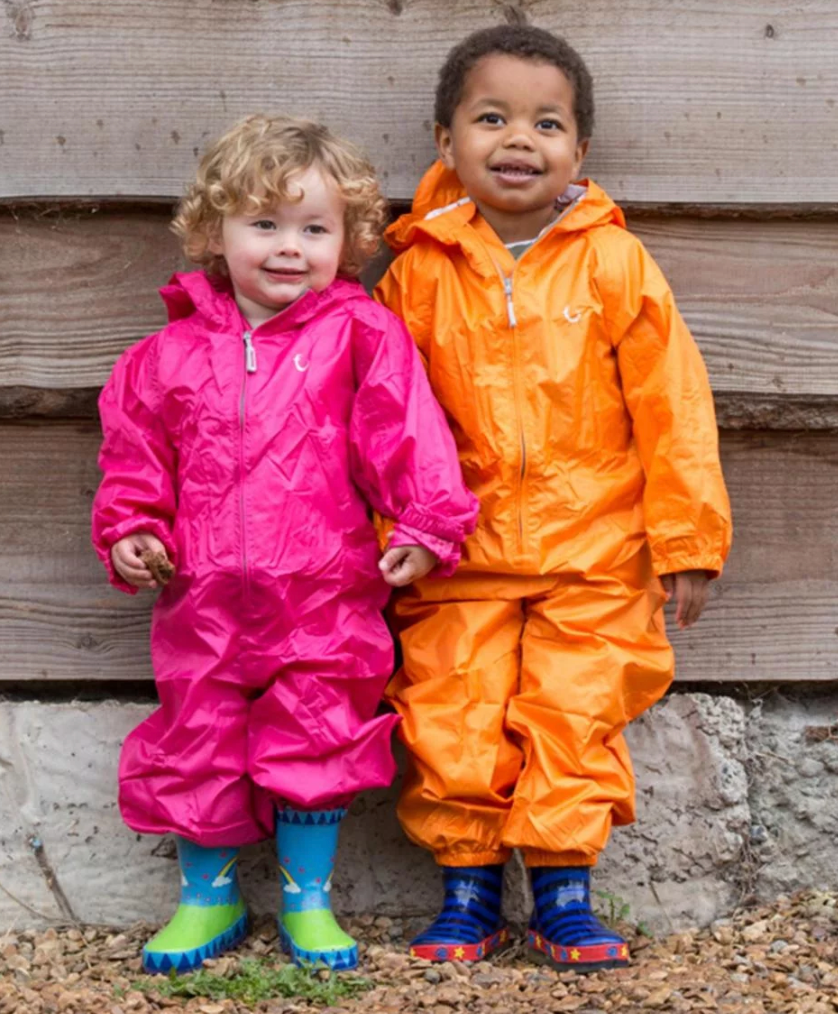 Two children wearing colorful rain suits standing against a wooden wall. WATERPROOF TODDLER ALL-IN-ONE PACKASUITS - PINK | HIPPYCHICK 