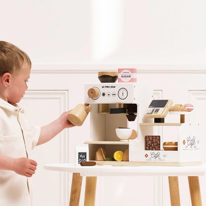 Child playing with a toy coffee set on a white surface. WOODEN BARISTA CAFE SHOP | LE TOY VAN