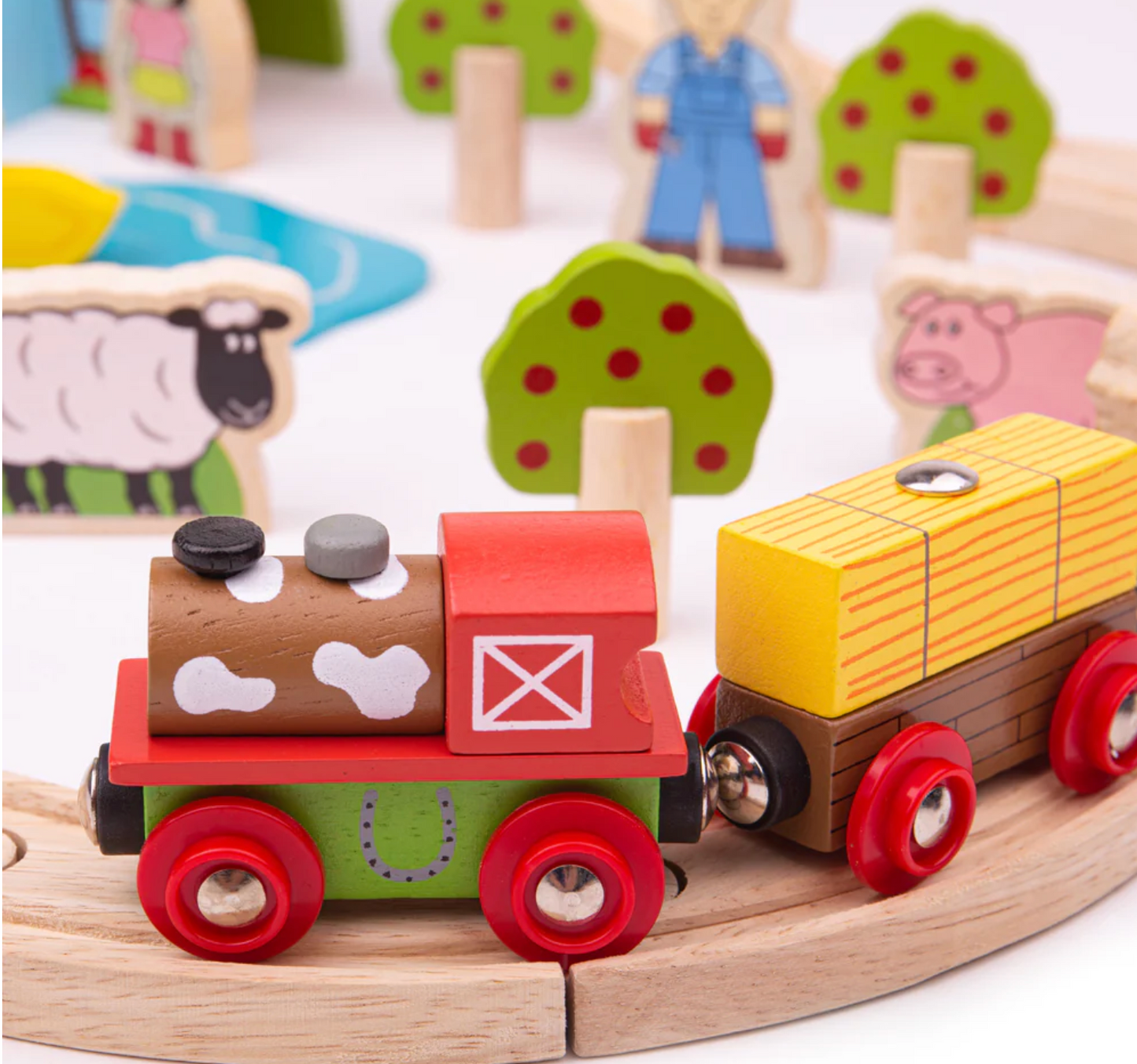 Wooden toy train set on a track with farm-themed figures and trees in the background. WOODEN FARM TRAIN SET | BIGJIGS